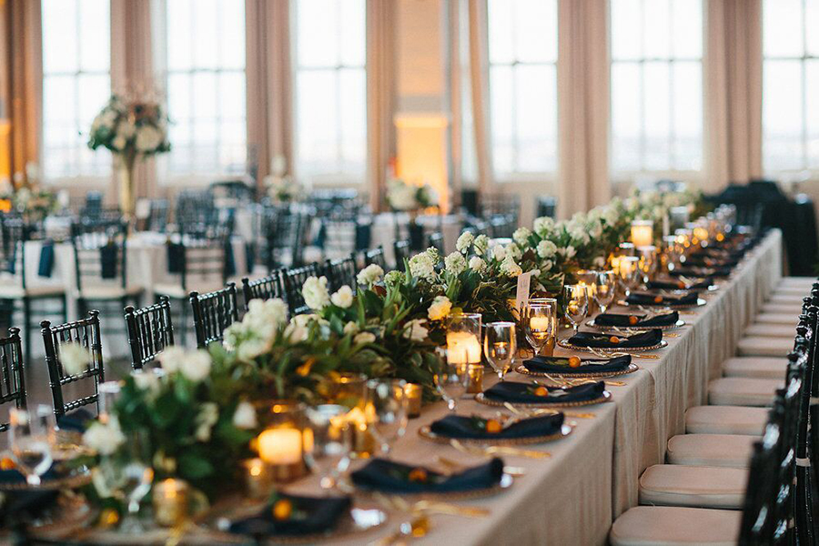 Long wedding reception head table with green and white floral garland, candles and placesetting with kumquats