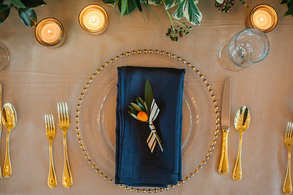 Wedding reception place setting with navy hanging napkin, gold-beaded charger, and kumquat detail