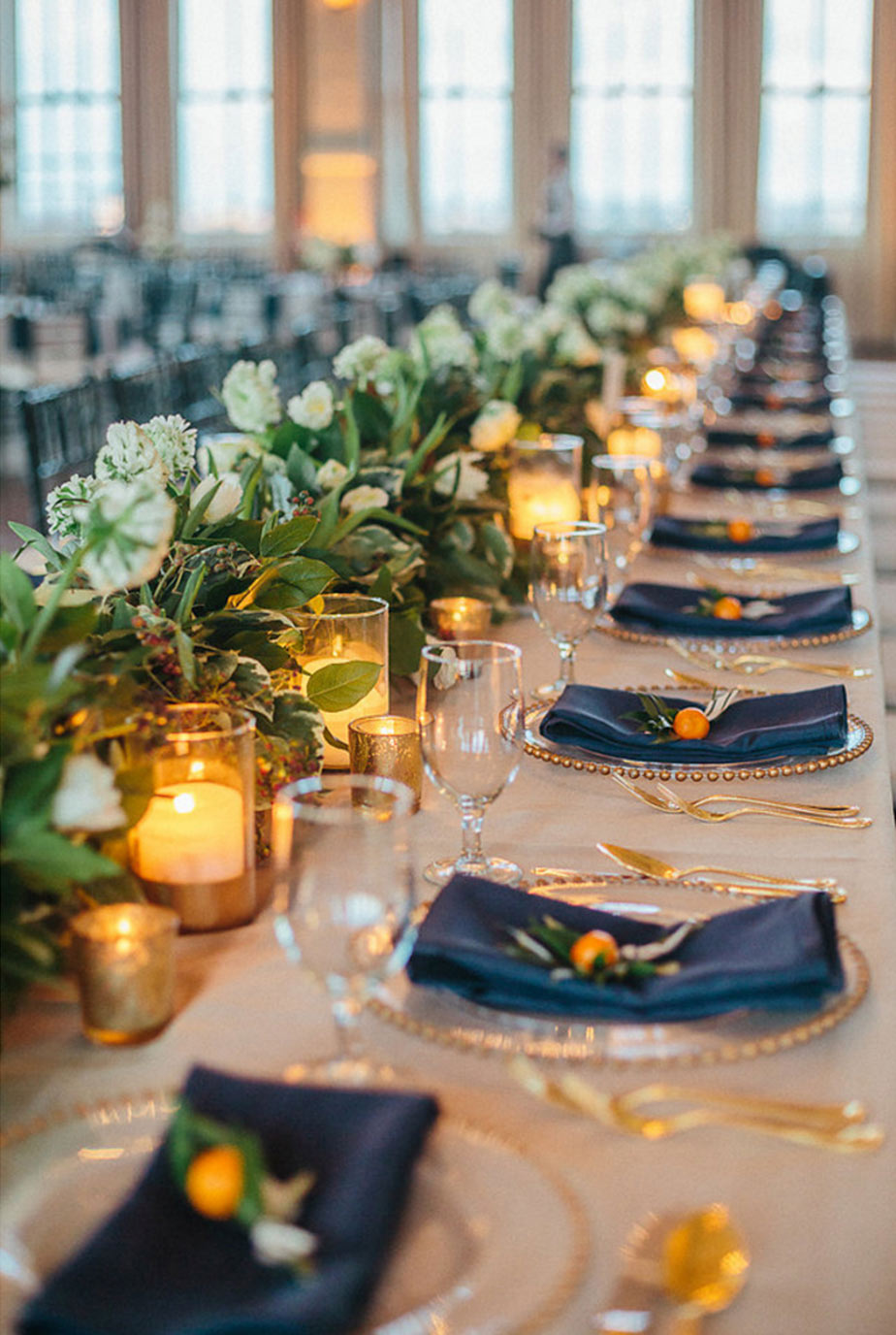 Long head table with floral garland and candles and place setting with navy hanging napkin, gold-beaded charger, and kumquat detail at wedding reception at The Room on Main in Dallas, Texas
