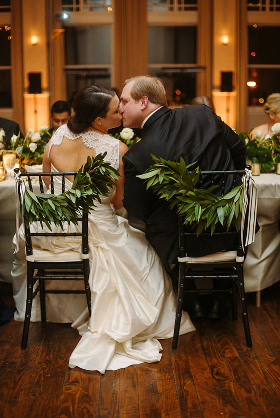 Bride and groom kissing at floral chair with swags of greenery and ribbon
