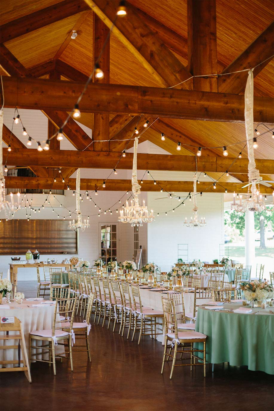 White Oaks Ranch wedding reception setup with gold chiavari chairs, chandeliers, wooden beams, and cafe lights