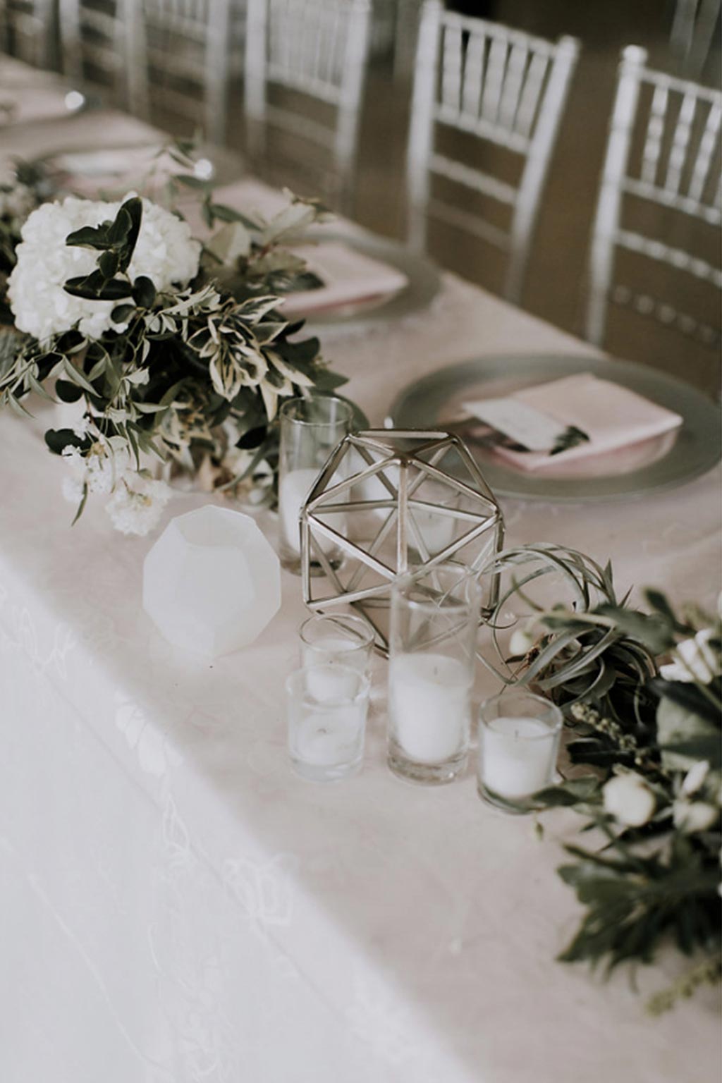 Long head table with silver chiavari chairs, faceted terrarium with green and white organic garland wedding centerpiece, and silver and glass, pink napkin placesetting with menu and fern leaf
